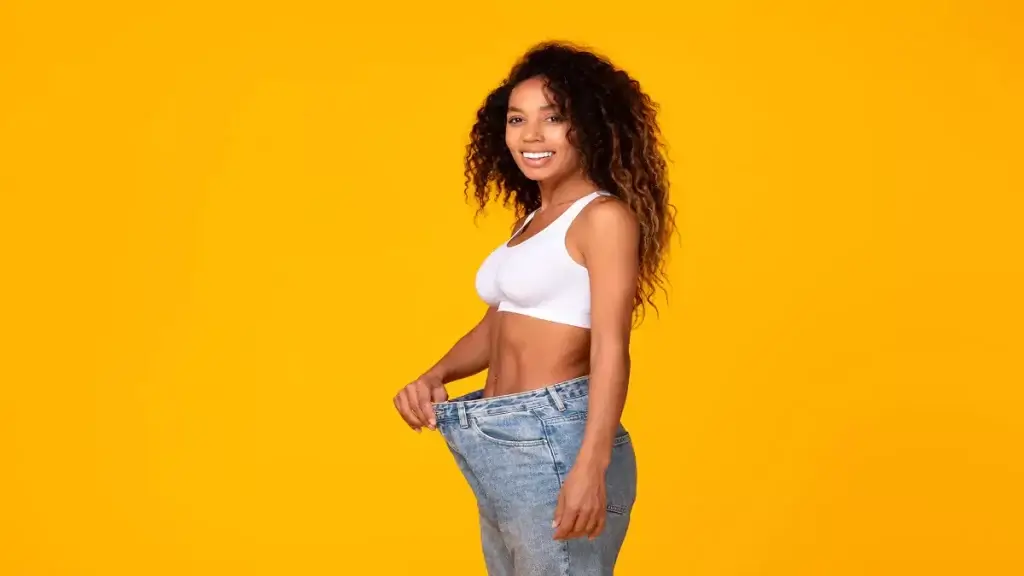 A woman is standing on a yellow background with her jeans on after liposuction.